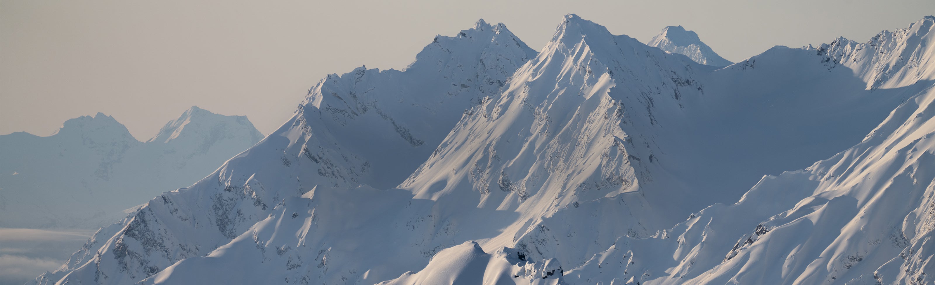 Snow-covered mountain peaks under a pale sky, with sharp ridges and broad white slopes.